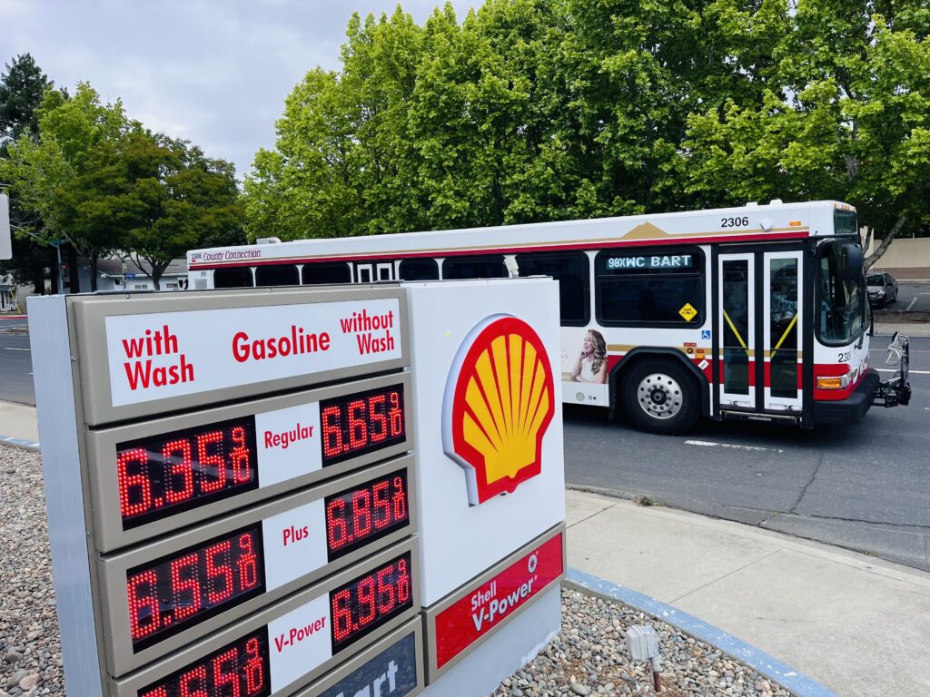Image of bus passing a gas station with price-per-gallon displayed in the foreground.